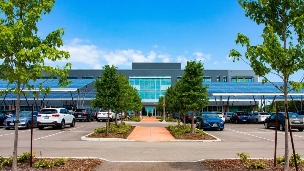 A photo of a parking lot with solar panels. An office building is in the background.